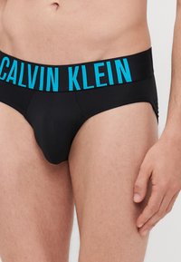 Schwarze Slips mit einem breiten elastischen Bund, auf dem "CALVIN KLEIN" in fetter blauer Schrift entlang des Taillenbands steht. Glatte Stoffoberfläche.
