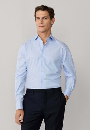 Man met een lichtblauw overhemd met lange mouwen en donkere broek, staand met één hand in de zak tegen een effen achtergrond.