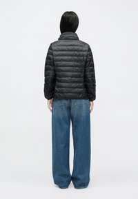 Melnā pārklātā dūnu jaka ar stāvošu apkakli un horizontālām šuvēm, kombinēta ar plašām zilām džinsa biksēm. Skats no aizmugures.