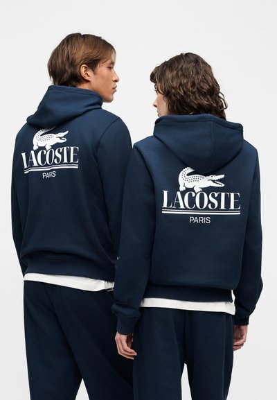 Двама души, облечени с тъмносини суичъри Lacoste с бели лога на крокодил на Lacoste и надпис "PARIS" на гърба, стоящи рамо до рамо с гръб към нас.