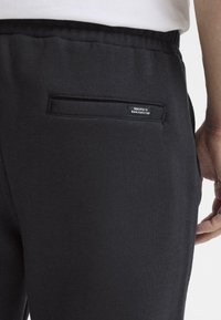 Czarne spodnie dresowe z tylną kieszenią i małą metką. Materiał ma gładką fakturę, a design jest prosty z elastycznym paskiem w talii.