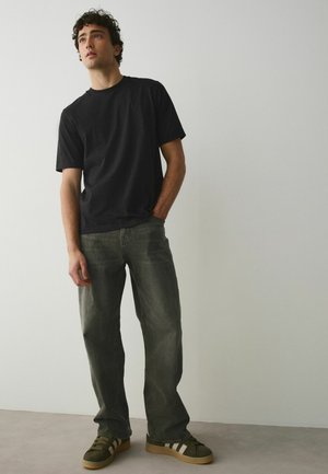 Fiatal férfi áll, fekete pólót, kopott olívazöld farmert és fehér csíkos zöld sportcipőt visel, egyszínű világos fal előtt.