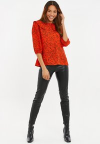 Rode blouse met zwart gespikkeld patroon, gerimpelde halslijn en pofmouwen, gecombineerd met zwarte glanzende fitted broeken en haklaarzen.
