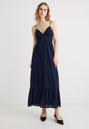 Kvinde iført en ærmeløs navyblå maxikjole med tynde stropper og sorte spidse hæle, stående foran en ensfarvet hvid baggrund.