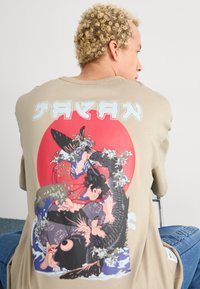 Persona con cabello rizado y rubio que lleva una camisa beige con una colorida obra de arte de estilo japonés que muestra a un guerrero luchando contra un pez grande.