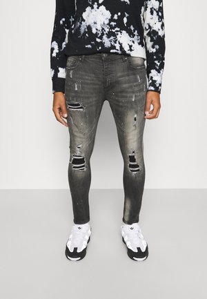 Persoon draagt zwarte en grijze versleten skinny jeans met scheuren bij de knieën en dijen, gecombineerd met witte en zwarte sneakers en een zwart tie-dye topje.