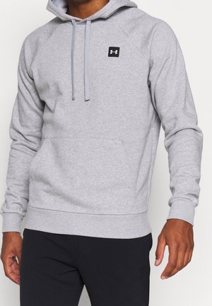 Mann trägt hellgrauen Under Armour Kapuzenpullover mit Fronttasche und schwarze Hose, steht vor einem schlichten hellen Hintergrund.