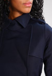 Kvinna med lockigt svart hår som bär en mörk marinblå kappa med bred krage och axelklaffar, läppar synliga, med ansiktet vänd mot vänster.