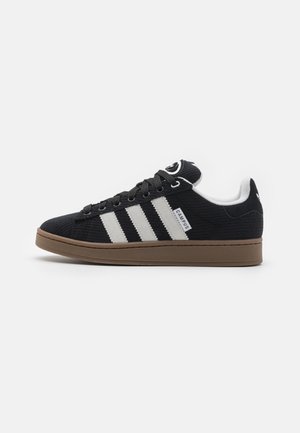 Sneaker en velours côtelé noir avec trois rayures blanches, une semelle en caoutchouc marron et un logo noir sur la languette. Tissu texturé avec un col rembourré.