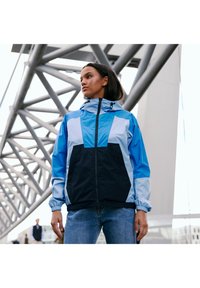 Windjack met een capuchon, featuring een kleurblokontwerp in lichtblauw, donkerblauw en wit. Elastiche manchetten en ritssluiting aan de voorkant.