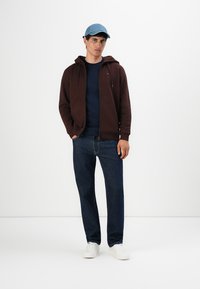 Joven con una gorra azul, sudadera marrón con cremallera, camisa azul marino, jeans azul oscuro y zapatillas blancas de pie frente a un fondo blanco.