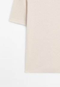 Texture de tissu beige de t-shirt à manches courtes montrée de côté avec des coutures visibles sur la manche et l'ourlet du bas.