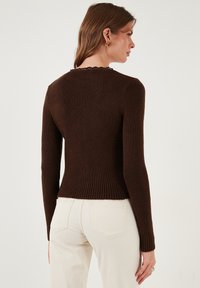 Bruine geribde gebreide trui met een golvende halslijn en lange mouwen; getailleerd ontwerp dat versmalt bij de taille. Draagt met lichte jeans.