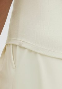 Krémovo sfarbený rebrovaný top s zaobleným lemom, s diskrétnou značkovou nášivkou na spodnej časti. Mäkka textúra, v kombinácii s hladkými svetlými nohavicami.