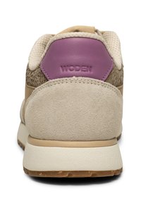 Beige sportssko med en texturerad tan mockaskal, lila hälaccent och vit mellansula; har vävda detaljer bak.