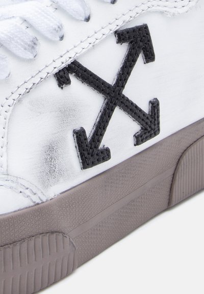 Baskets blanches avec un logo "X" noir cousu. Semelle en caoutchouc texturée et lacets. Le matériau semble lisse avec de légères marques d'usure.