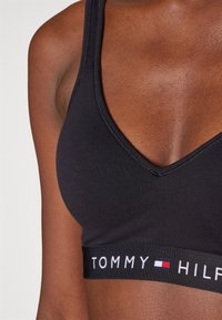 Svart sport-bh med djup V-ringning, tillverkad av mjukt tyg. Har en vit "TOMMY HILFIGER"-logotyp på ett texturerat band vid botten.