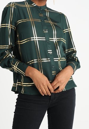 Woman wearing a dark green long-sleeve buttoned shirt with gold and white plaid pattern, paired with black pants. Hands clasped at waist.