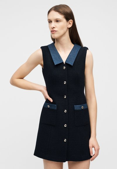 Γυναίκα που φοράει μαύρο φόρεμα χωρίς μανίκια με μπλε γιακά και καλύμματα τσεπών, με κούμπωμα μπροστά με κουμπιά, στέκεται μπροστά σε ένα απλό γκρι φόντο.