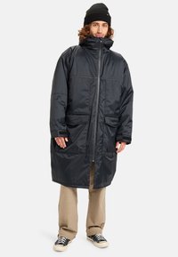 Zwarte nylon parka-jas met hoge kraag, ritssluiting, twee voorkruiszakken, lange mouwen en een relaxte pasvorm; gedragen over lichtgekleurde broeken.