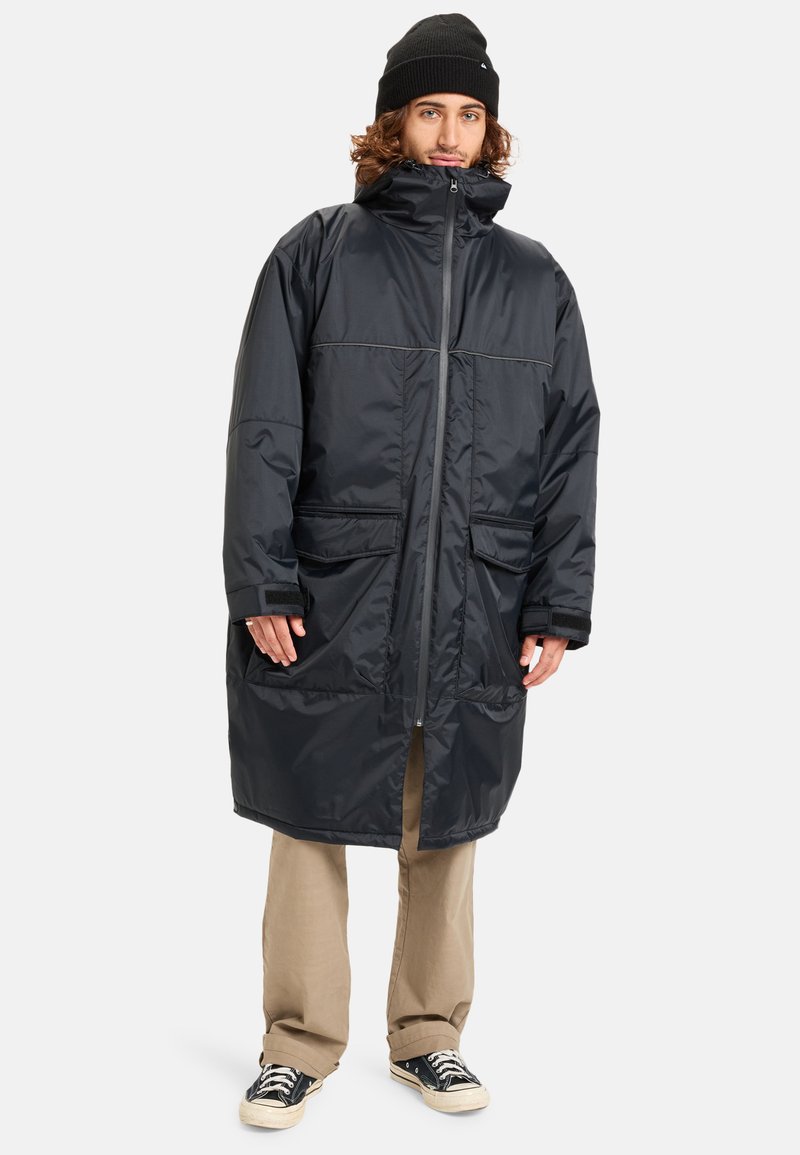 Zwarte nylon parka-jas met hoge kraag, ritssluiting, twee voorkruiszakken, lange mouwen en een relaxte pasvorm; gedragen over lichtgekleurde broeken.