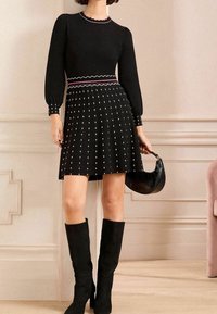 Žena v černých dlouhých šatech s bílou puntíkovanou sukní, černých kozačkách po kolena, držící černou kabelku v béžovém pokoji.