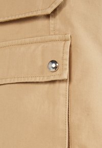 Brun tyg med en synlig, strukturerad yta. Har en framficka med en silverknapp som detalj och markerad sömnad längs kanterna.