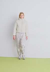 Giacca imbottita grigia chiara con colletto alto e zip anteriore abbinata a pantaloni larghi grigi chiari con pattern e sneakers metallizzate.