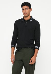 Melnas garām piedurknēm polo krekls ar baltām svītrām apkaklē un piedurknēm, ar izšūtu logo uz krūšu daļas. Saskaņots ar tumši zaļām biksēm.