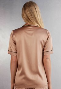 Satin pyjamas topp i ljusbrun med korta ärmar och svarta detaljer. Slät yta och skräddarsydd krage. Bakvy som visar designen.