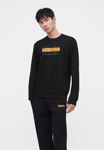 Мъж, носещ черна суитчерка EA7 Emporio Armani и черни панталони, стоящ срещу обикновен светъл фон.