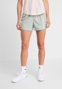 Mujer vistiendo pantalones cortos deportivos verde claro, calcetas blancas de Nike y zapatillas blancas, de pie contra un fondo blanco liso.