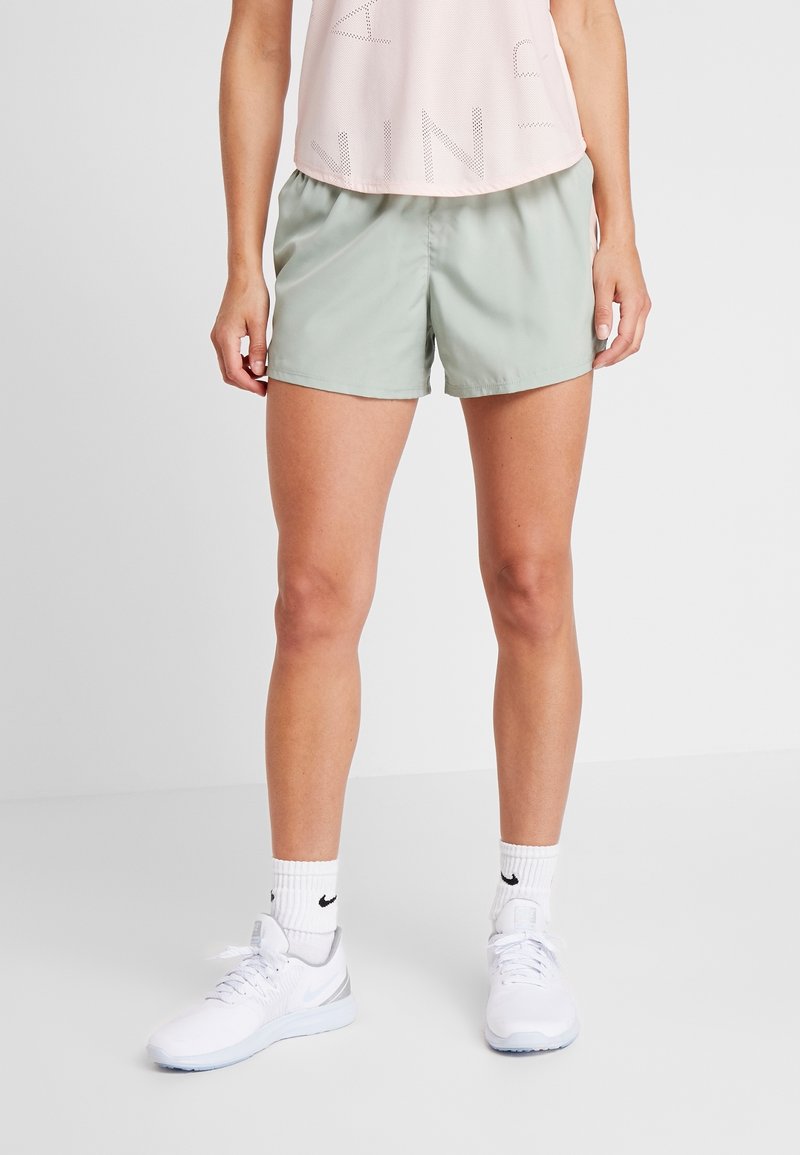 Mujer vistiendo pantalones cortos deportivos verde claro, calcetas blancas de Nike y zapatillas blancas, de pie contra un fondo blanco liso.