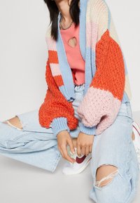 Cárdigan de punto extragrande multicolor con rayas en naranja, rosa, azul y crema. Combinado con una blusa rosa, jeans claros desgastados y zapatillas blancas.