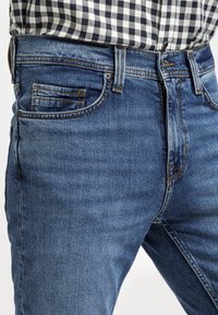 Niebieskie dżinsy o prostym kroju, pięć kieszeni i kontrastowe przeszycia. Noszone z czarno-białą koszulą w kratę.