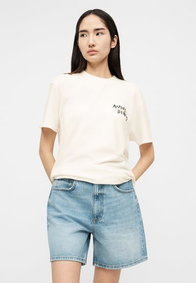 Woman wearing a white graphic t-shirt and light blue denim shorts standing with hands in pockets, looking to the side on plain background.