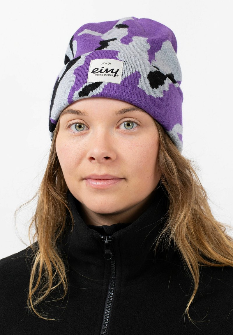 Purple and gray patterned knit beanie with a visible brand logo patch, worn above a black zippered fleece jacket. Soft and textured material.