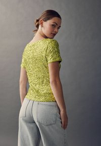 Glittrande gul paljett-topp med korta ärmar, kombinerad med ljusgrå högmidjade jeans. Minimalistisk design.