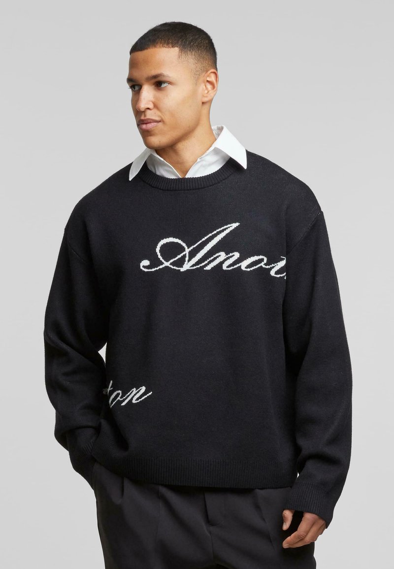 Suéter negro de punto con texto en cursiva blanca. Tiene un cuello redondo, hombros caídos y puños y dobladillo acanalados para dar textura.