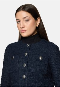 Femme aux longs cheveux bruns portant une veste texturée bleu foncé avec des boutons argentés sur un col roulé noir, posant devant un fond uni.