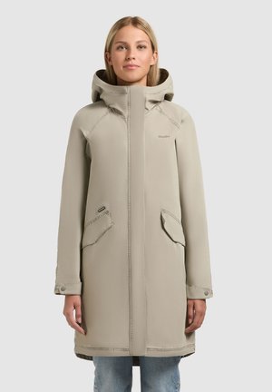 Vrouw die een beige gewatteerde jas met capuchon en voorzakken draagt, staand tegen een effen lichtgrijze achtergrond.