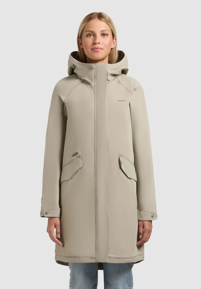 Femeie purtând o haină bej cu glugă și buzunare frontale cu clapetă, stând pe un fundal simplu gri deschis.