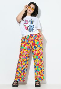 Pantaloni stampati colorati con motivi di gelato e frutta; abbinati a una t-shirt bianca con grafica; sandali neri con zeppa.