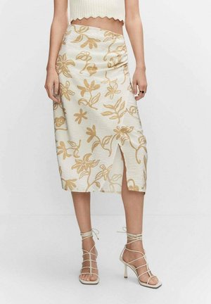 Woman wearing a white midi skirt with beige floral print and front slit, paired with white strappy high-heeled sandals and a cropped top.