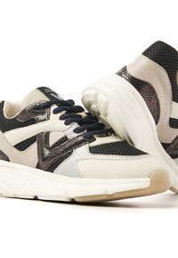 Ein Paar beiger, schwarzer und metallischer Sneaker mit strukturierten Details, marineblauen Schnürsenkeln und dicken weißen Sohlen, die sich auf einem weißen Hintergrund gegenüberstehen.