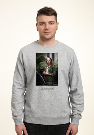 Mężczyzna w szarej bluzie z kapturem z zdjęciem kobiety trzymającej czerwony folder na zewnątrz, stoi obok czarnego samochodu, poniżej tekst „gossip girl“.