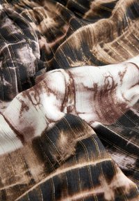 Tkanina prikazuje tie-dye uzorak u zemljanim tonovima crne, smeđe i krem, s metalik zlatnim linijama koje poboljšavaju njenu teksturu i dizajn.