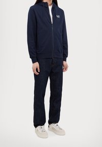 Marinblå zip-up-hoodie med kenguruficka och dragsko i huvan, kombinerad med mörka denimjeans och ljusfärgade sneakers.