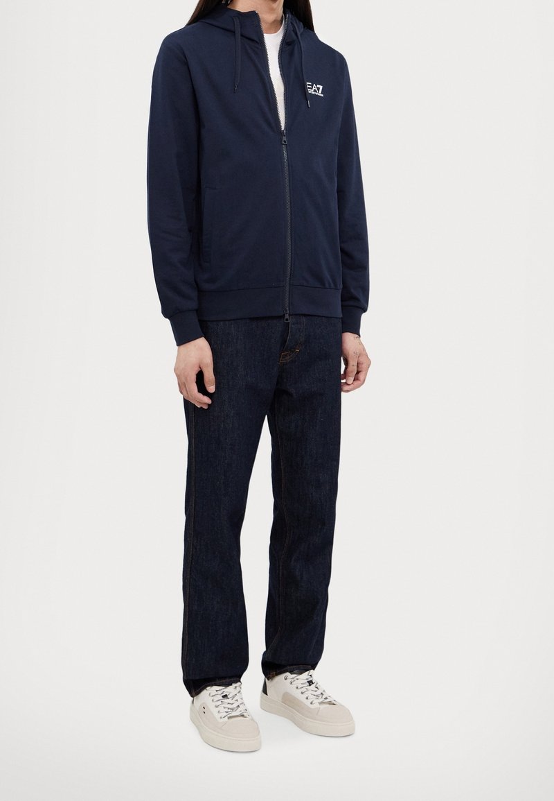 Marinblå zip-up-hoodie med kenguruficka och dragsko i huvan, kombinerad med mörka denimjeans och ljusfärgade sneakers.