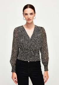 Blusa nera con pois bianchi, scollatura a V profonda, maniche lunghe trasparenti, vita arricciata e tessuto smock. Abbinata a jeans neri.